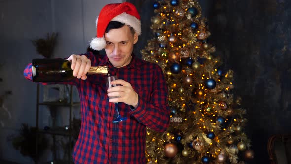 Young Man in Santa Claus Hat Pours Champagne Into Glass Standing Near Christmas Tree