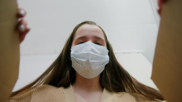 Young Woman During Pandemic Wearing Medical Face Mask Opening Cardboard Box with Order From Internet