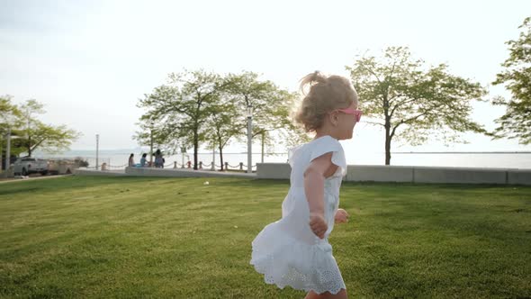 Little Girl in a White Dress Runs in the Park on the Green Grass on a Sunny Day