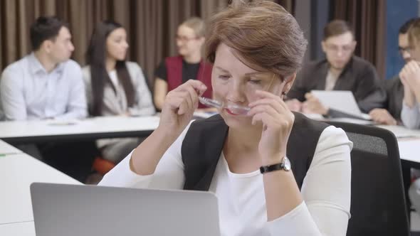 Serious Confident Woman Putting on Eyeglasses and Messaging Online on Laptop with Group of Blurred