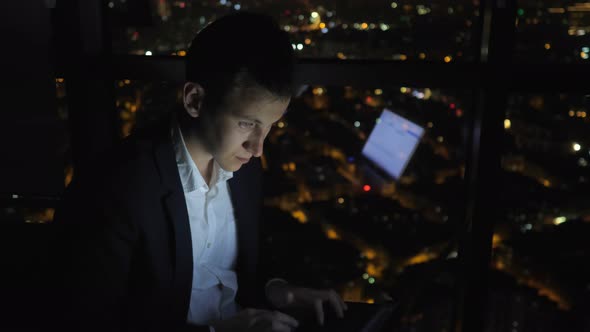 Young Man Freelancer Is Working on His Computer Near the Window in Home at Night