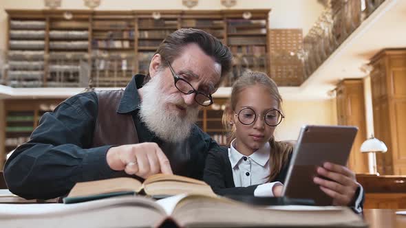 Bearded Grandpa Sitting at the Table in Library with His Pretty Granddaughter