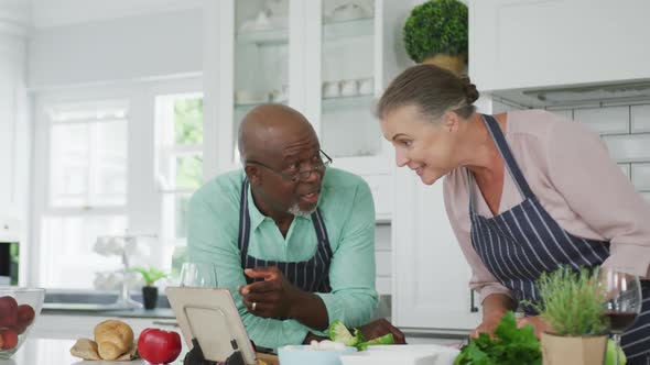 Smiling senior diverse couple wearing blue aprons and using tablet in kitchen