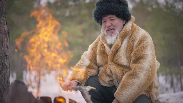 Senior Indigenous Man Looking at Fire Burning in Slow Motion Smiling Sitting in Winter Forest