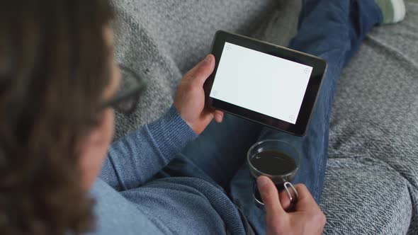 Caucasian man sitting on sofa in living room using tablet and drinking coffee