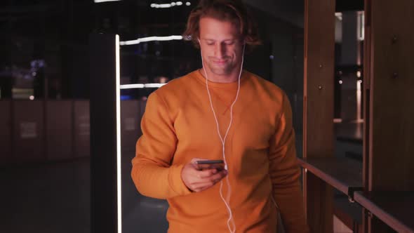 Caucasian male wearing earphones and skateboard, smiling, walking, using his phone in the evening