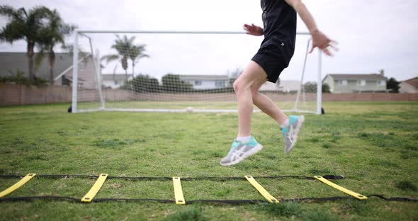 A young athletic girl practices high knees on a speed ladder at the park with a soccer goal in the b