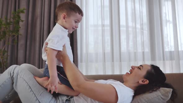 Happy Healthy Family Young Mom Lifting Cute Little Child Son Up Playing Plane on Sofa at Home, Funny