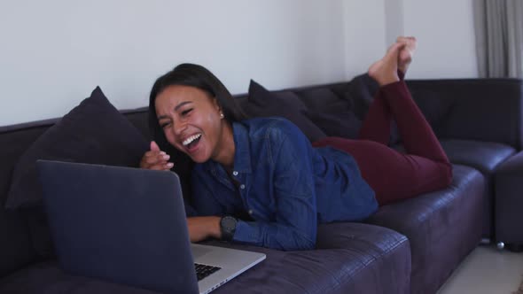 Mixed race woman lying on couch using laptop and laughing