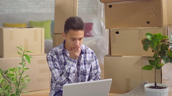 Young Thoughtful Asian Man After Moving To a New Apartment Sitting at a Laptop on the Floor