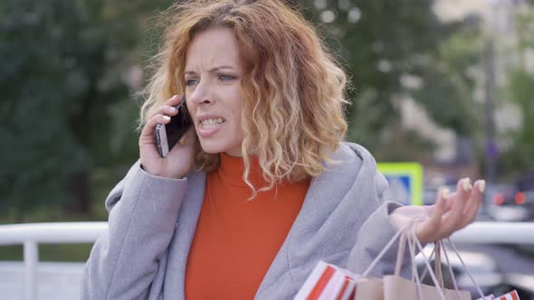 Unsatisfied Red-haired Woman with Shopping Bags Talking By Her Cellphone. Shopaholic Standing with