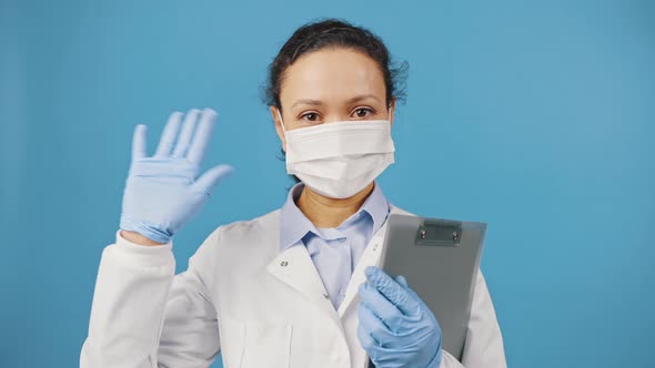 Friendly Woman Doctor Wearing Protective Face Mask and Holding Clipboard Waving Hand Hello to Camera