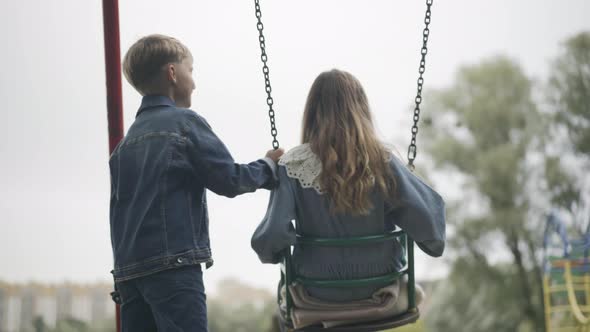 Back View of Happy Relaxed Girl Sitting on Swings As Loving Cute Boy Swinging Her