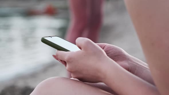 Beautiful Girl in a Bathing Suit Sits on a Sandy Beach with a Phone in Her Hands