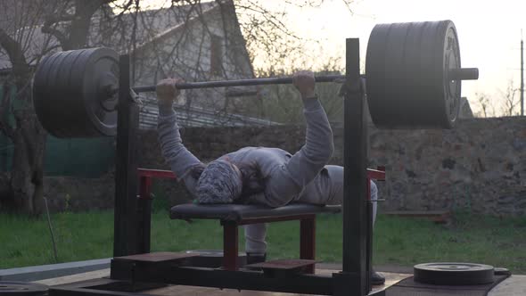 A Man an Athlete is Training Outdoors Doing Chest Press with a Barbell