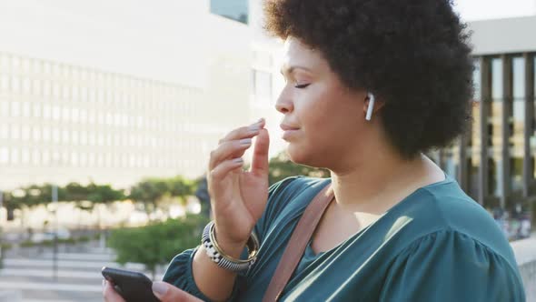 Happy plus size biracial woman using smartphone and wearing earphones in city