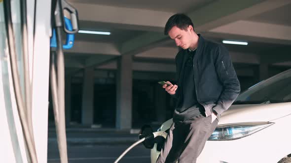 Man using smartphone and staying near his charging electric car