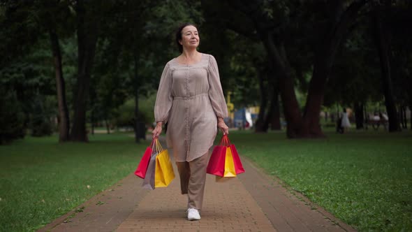 Wide Shot Portrait Gorgeous Confident Senior Woman Walking with Shopping Bags in Park Smiling