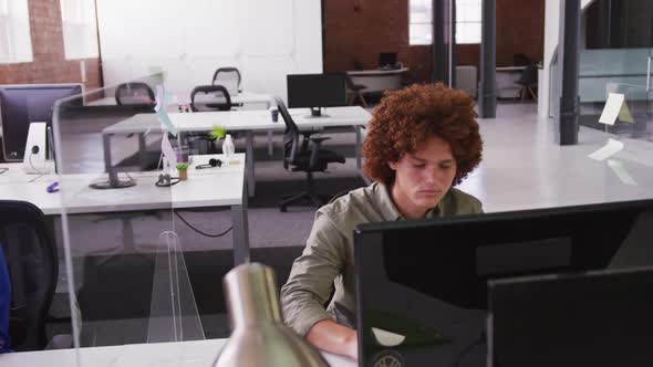 Mixed race businessman sitting at desk with sneeze guard using desktop computer looking at screen