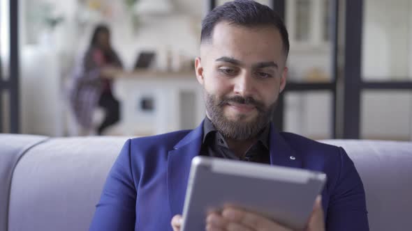 Attractive Bearded Man in Blue Suit Sitting at Home Working with His Tablet. Leisure Indoors. The