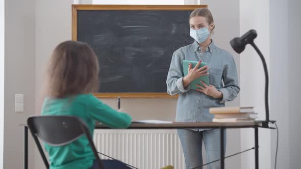 Portrait of Intelligent Teacher in Coronavirus Face Mask Talking with Student Raising Hand Asking