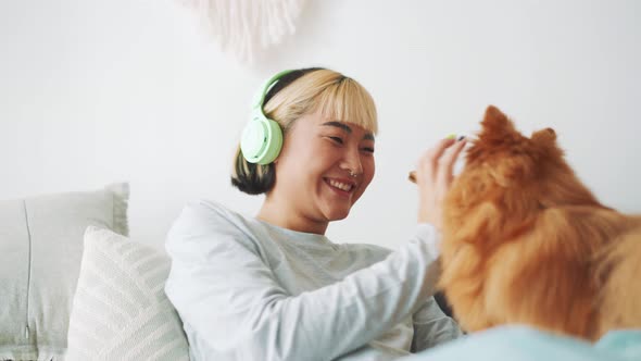 Laughing Asian woman in headphones stroking her dog on the bed