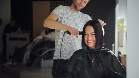 Male Professional Hairdresser Is Straightening Brunette Woman's Hair Using a Hairstraightener in