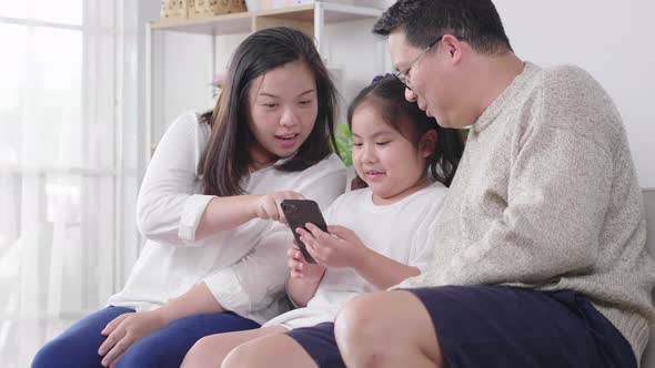 Happy asian family parent and daughter having fun with gadget on couch using smartphone at home
