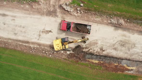 Top View of a Yellow Excavator Loads Soil From the Field Into a Red Truck During Excavation Work