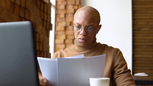 An African American Man with a Serious Expression Works with a Laptop and Documents at Home