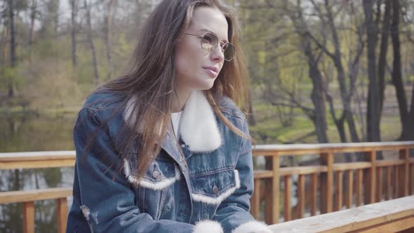 Portrait of Young Glamour Woman in Sunglasses and Jeans Jacket Standing on the Wooden Bridge Looking