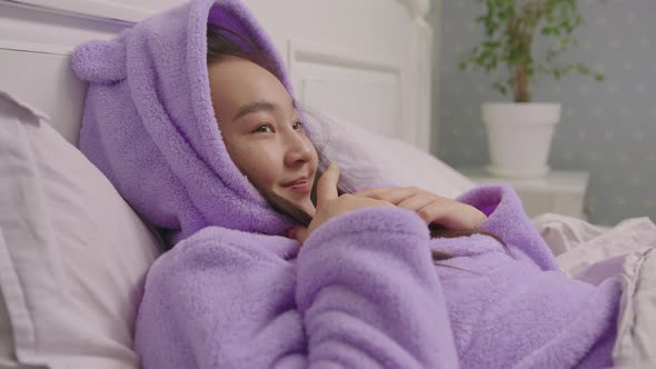 Happy 20s Asian Woman in Purple Pajamas Lying in Bed and Covering Herself By Duvet Blanket