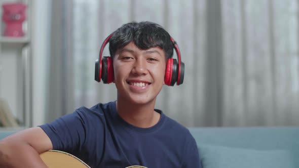 Asian Boy Composer Wearing Headphones And Smiling To Camera While Playing Guitar At Home