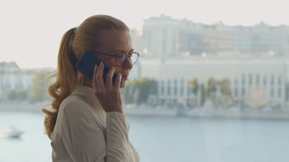 Mature Businesswoman in Glasses Talking on Mobile Phone Looking Out Window in Business Center