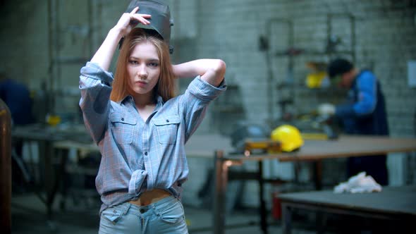 Young Sexy Woman in Small Shorts Standing in the Workshop and Putting on Her Helmet - Man Working on