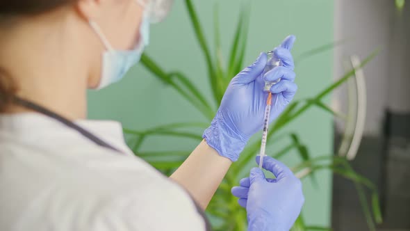 A Doctor in a Medical Mask and Blue Gloves Holds a Syringe and a Vaccine