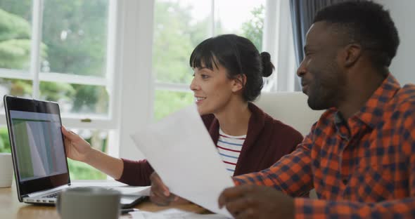 Happy diverse couple sitting at table talking and working with laptop