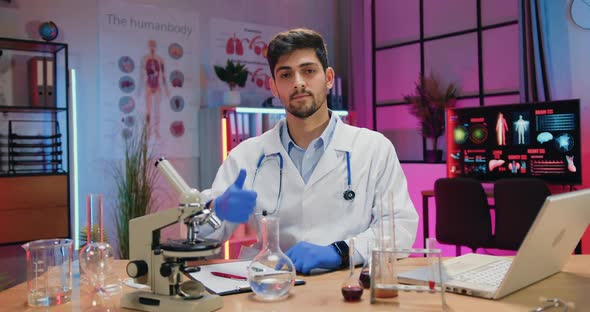 Chemist in Medical Uniform and Gloves Sitting in front of Camera in Evening Lab