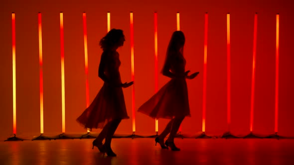 Two Graceful Young Women Practice the Elements of Salsa Dance in a Dark Studio Against a Background