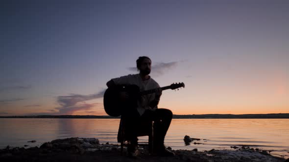 Silhouette of a Man Playing Guitar Sitting on a Chair By the Lake on Colourful Sunset Background