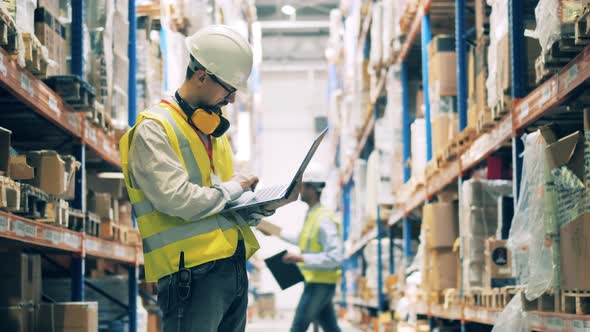 Storeman in Uniform Typing on Laptop Among Warehouse Shelves