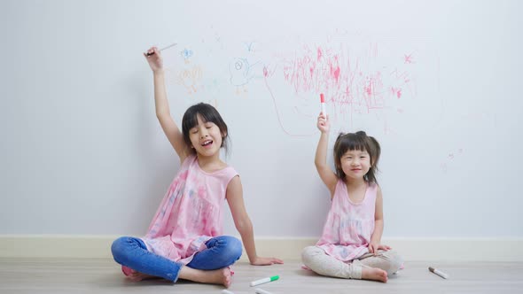 Asian young sibling kid girl having fun drawing and coloring on white wall in living room at home.