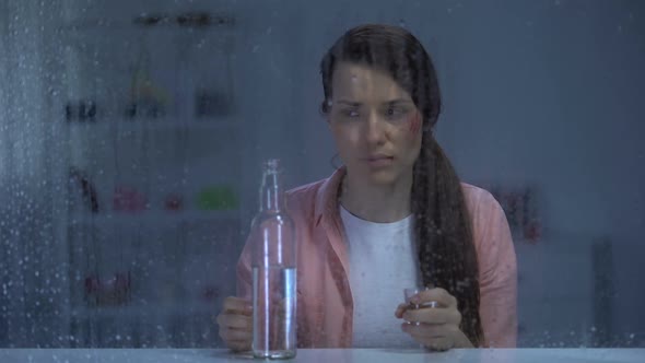 Sad Woman With Wounded Face Drinking Vodka Behind Rainy Window