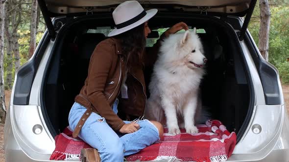 A young woman in a stylish hat sits with a dog in the trunk, in the woods. Traveling with animals.