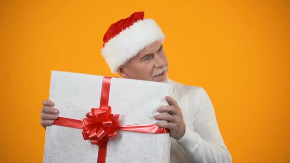 Positive Male Pensioner in Santa Claus Hat Shaking Giftbox and Smiling, Surprise