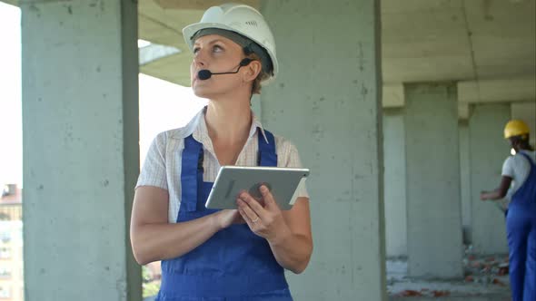 Female Construction Engineer Reading Plans Using Digital Tablet and Talk To Workers Through Internal alt