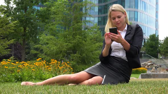 A Young Beautiful Businesswoman Sits on Grass in a Park and Works on a Smartphone - Office Buildings