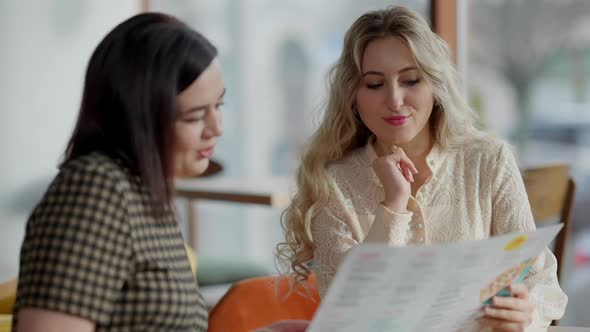 Two Positive Young Caucasian Women Choosing Food in Restaurant Menu