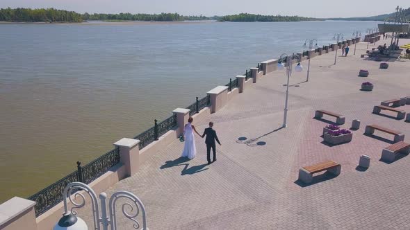 Just Married Couple Walks Along Waterfront Near River Aerial