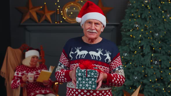 Elderly Grandfather in Festive Sweater Presenting Christmas Gift Box Smiling Looking at Camera